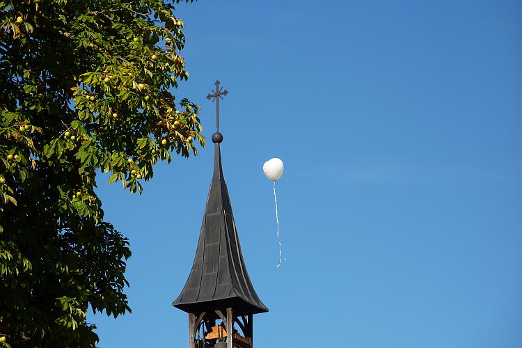 weißer Luftballon in Herzform neben Glockenturm eines Kirchleins vorbeifliegend