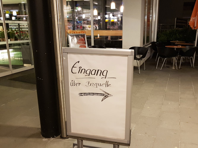 Plakatständer vor dem MPreis, Text "Eingang über Baguette", der Pfeil darunter zeigt in die falsche Richtung